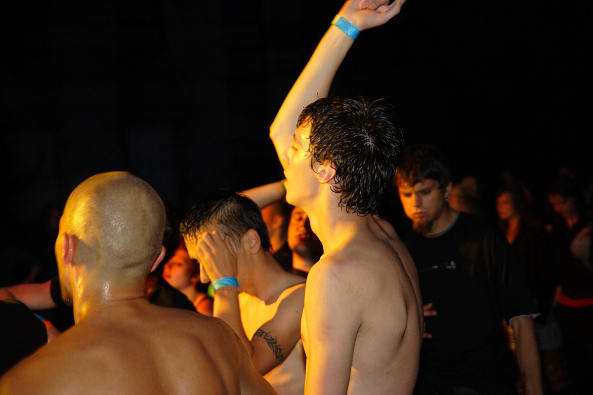 A group of people at a 2009 metal show, illuminated by warm lighting. In the foreground, a shirtless man with wet hair raises his arm energetically. Another man with a shaved head is nearby. The background is filled with people, some facing forward and others turned away.