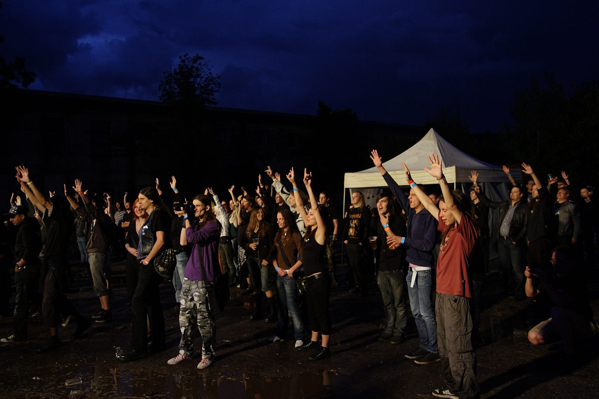 A group of people stands outdoors in front of a white tent at night, raising their hands enthusiastically. The sky is dark and stormy, creating a dramatic atmosphere. Most individuals appear engaged and attentive, suggesting they might be watching a performance at the Metalshow '09.