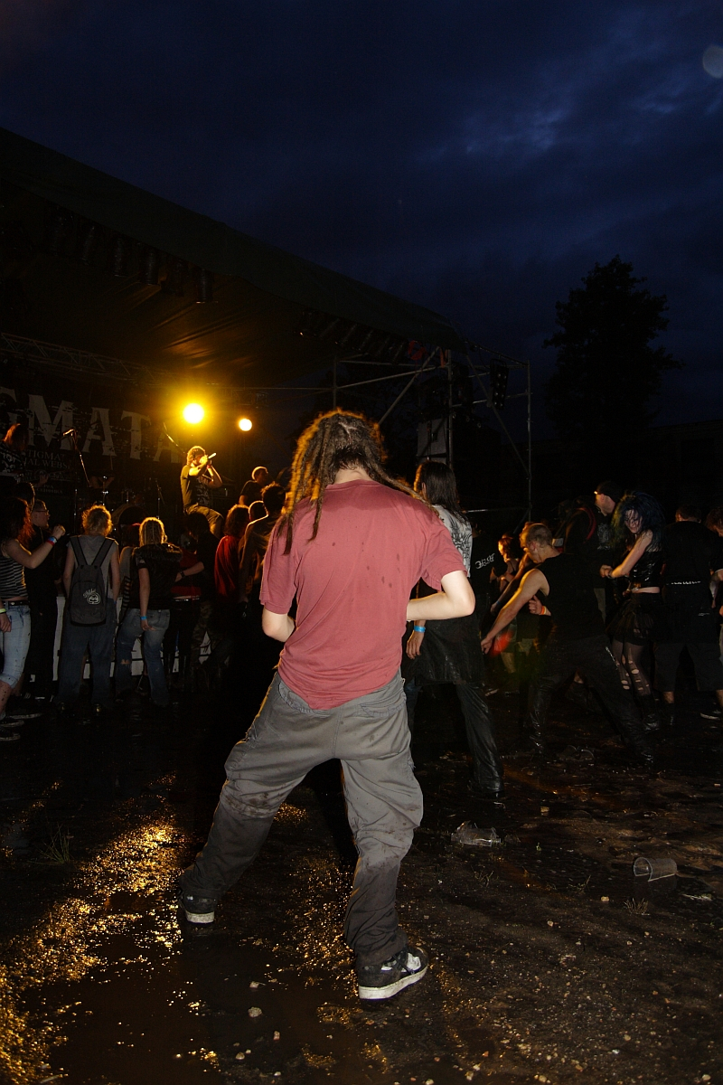 A person with dreadlocks stands in a wet, muddy area at an outdoor concert during nighttime. They wear a red shirt and gray pants. In the background, a crowd of people watches a performance on stage, illuminated by bright yellow light. The sky is dark and cloudy, capturing the essence of the 2009 metal show event.