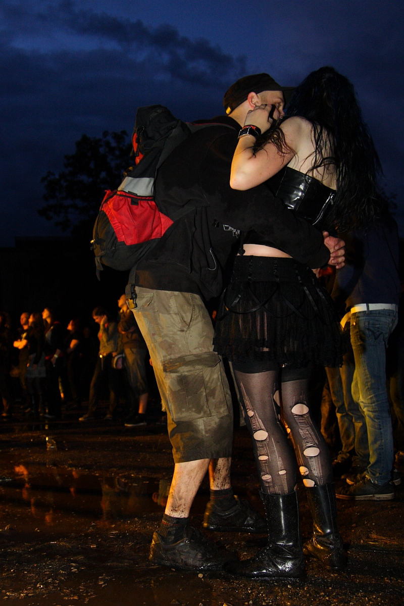 A couple stands closely, embracing each other at a metal event during dusk. The person on the left wears a black cap, black jacket, tan cargo shorts, and a black and red backpack. The other person is dressed in a black top, black skirt, ripped tights, and black knee-high boots. A crowd is visible in the background.