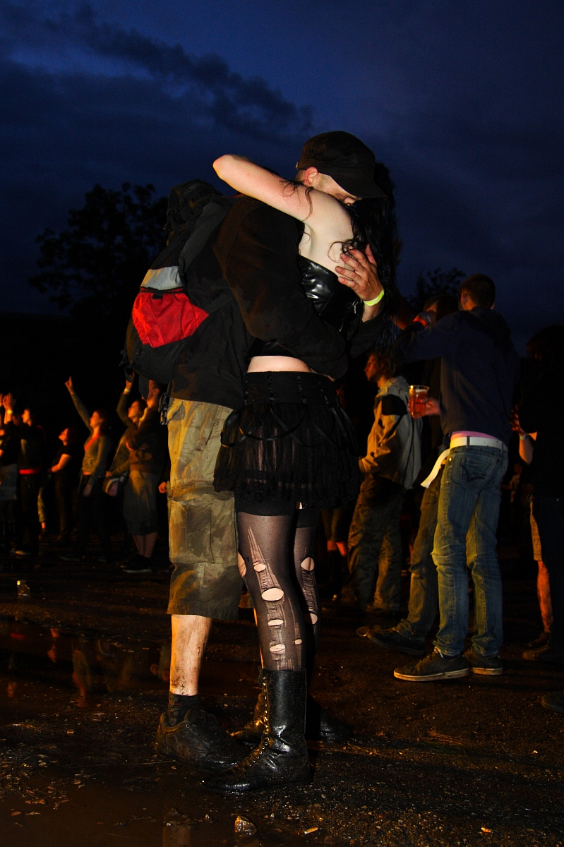 A couple embraces at an outdoor event during nighttime. The woman, dressed in dark clothing with patterned tights and boots, has her arms wrapped around a man wearing a cap, dark jacket, red backpack, and camouflage pants. People in the background of this Metalshow '09 are engaged in various activities.