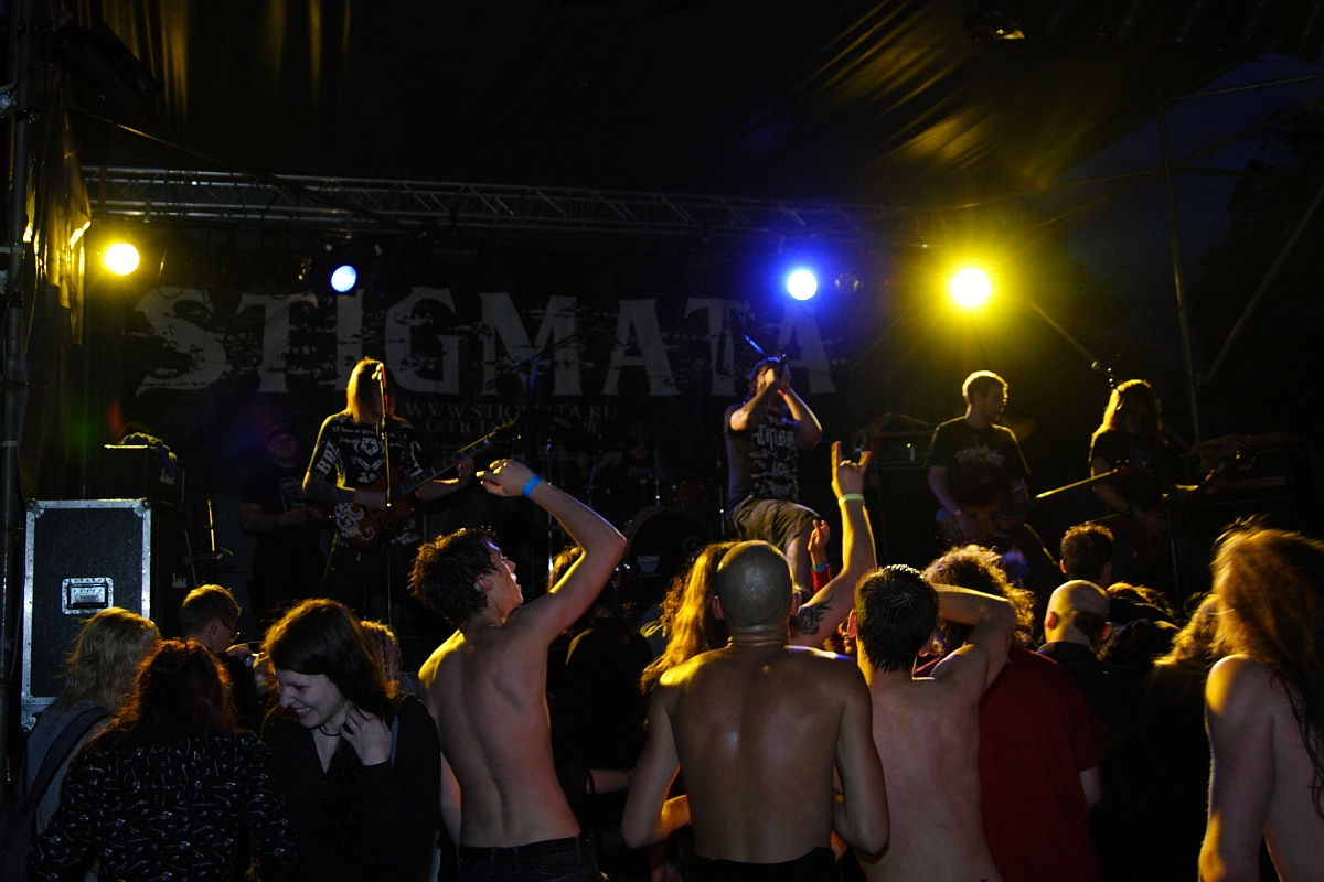 A live band performs energetically on stage under intense yellow and blue lighting at Metalshow '09. The audience, some shirtless and others clothed, enthusiastically raises their arms and cheers. The band's backdrop displays the word "STIGMA." The dark atmosphere highlights the lively concert vibes.