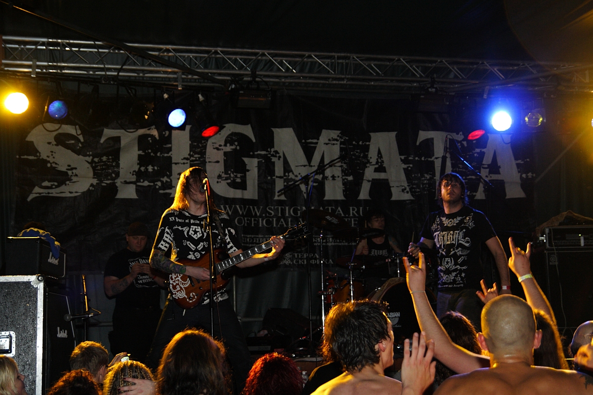 A rock band performs energetically on stage with a large "Stigmata" banner behind them at a 2009 metalshow. The band members wear dark, graphic clothing and play electric guitars and drums. The audience, some with raised hands, enjoys the lively atmosphere under colorful stage lights.