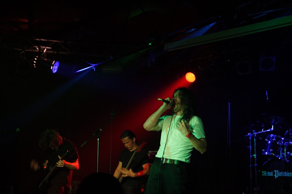 A band performing on a dark stage with dramatic lighting. The lead singer, wearing a white shirt, is holding a microphone and singing passionately at this 2009 metalshow. Two guitarists are on either side, focused on their instruments. In the background, a drum set is partially visible under dim red and blue lights.