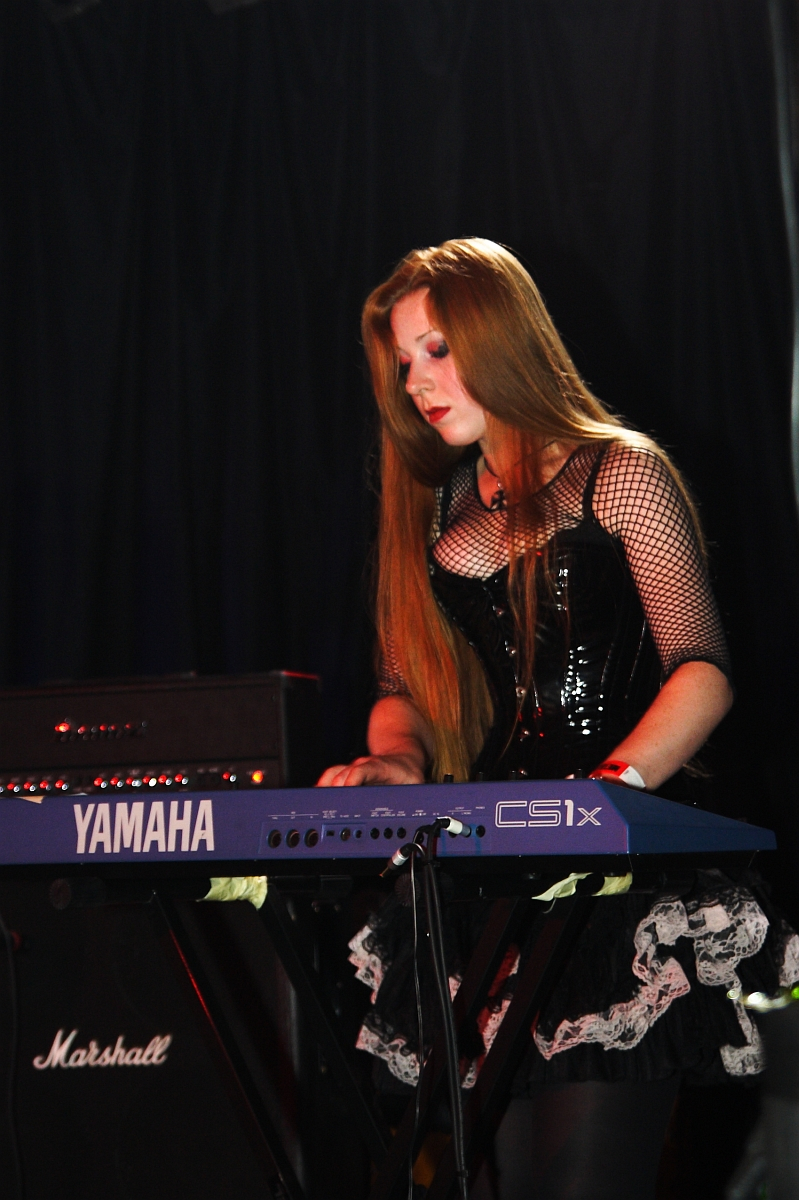 A woman with long red hair is playing a Yamaha CS1x keyboard on stage at Metalshow '09. She is wearing a black corset-style top with mesh sleeves and accessorized with a frilly skirt. A Marshall amplifier is positioned behind her against a backdrop of black curtains. The scene is dimly lit, creating a dramatic atmosphere.