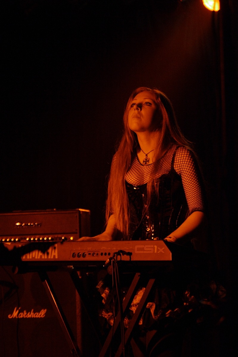 In a 2009 metalshow, a woman with long hair performs on a keyboard under dim, warmly colored stage lighting. Dressed in a black corset, mesh sleeves, and a skirt, she wears a cross necklace. Behind her is a Marshall amplifier. The scene is dark, focusing attention on the performer and her instrument.