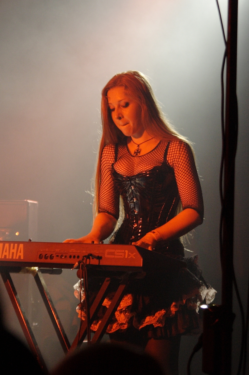 A woman with long hair is playing a Yamaha keyboard on stage at Metalshow '09. She is wearing a black corset dress with fishnet sleeves and a layered lace skirt. The background is smoky and dimly lit, creating a moody atmosphere. A microphone stand is partially visible to the right.