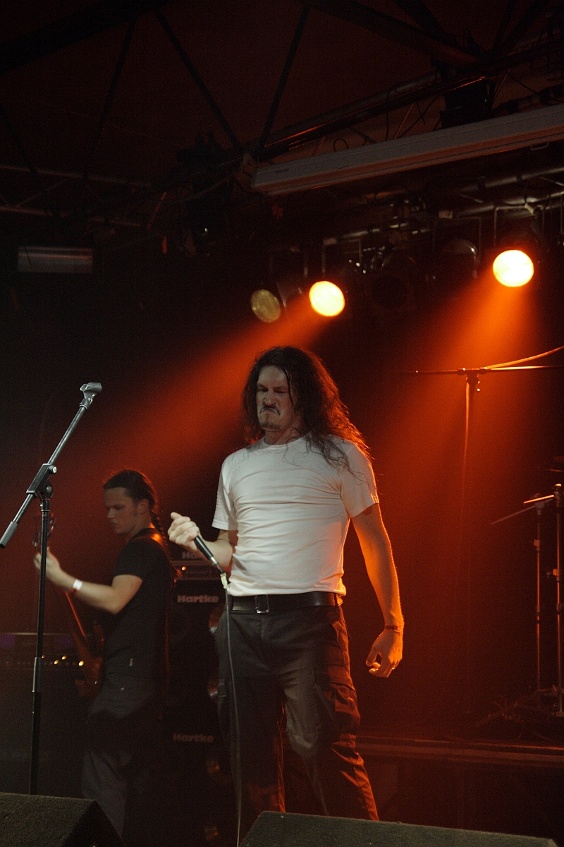 A long-haired man in a white shirt and dark pants passionately sings into a microphone on a dimly lit stage with orange lighting at Metalshow '09. Another musician in black stands behind him, playing a guitar. Music equipment and spotlights are visible in the background, adding to the intense atmosphere.
