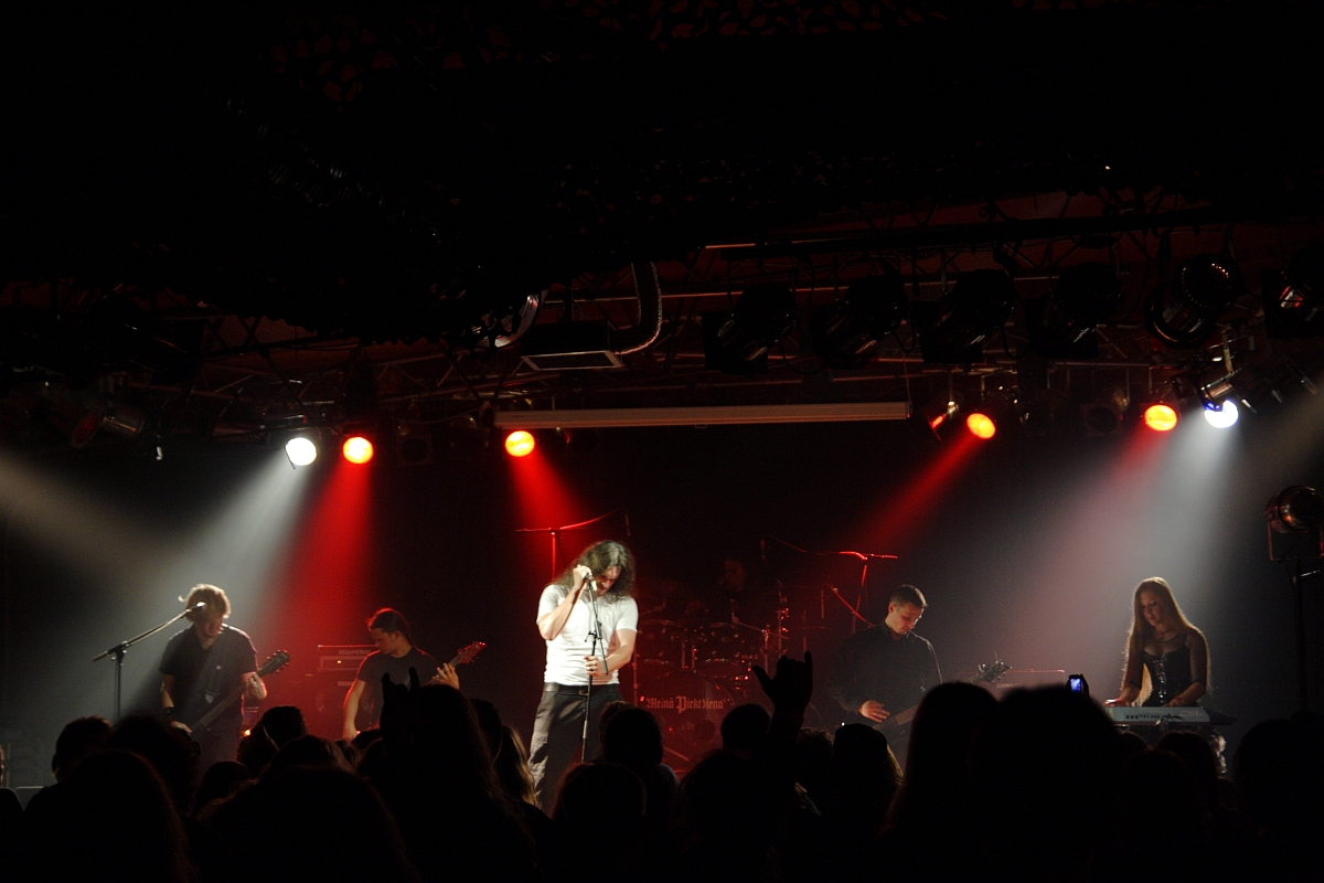 A rock band performing on stage with five members visible. The lead vocalist is in the center, singing passionately into a microphone. Other band members are playing guitars, a keyboard, and drums. The stage is lit with vibrant red and white lights, reminiscent of a 2009 metalshow, and an audience is silhouetted in the foreground.