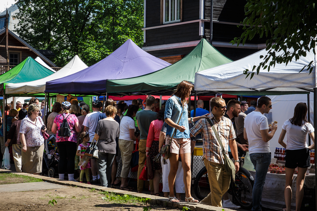 A bustling outdoor market with numerous people interacting under colorful tents at Kalnciema kvartāla. Some individuals are browsing, while others are engaged in conversation. The green, purple, blue, and white tents add to the vibrant scene of modes diena set against a backdrop of trees and buildings.