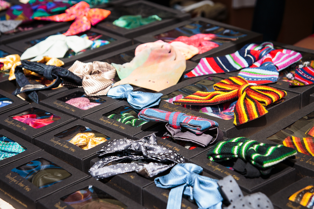 A display table at Kalnciema kvartāls is filled with variously colored and patterned bow ties, neatly arranged in black boxes with clear plastic windows. The ties have designs ranging from polka dots and stripes to solid colors in bright and pastel hues, perfect for modes diena in Latvija.