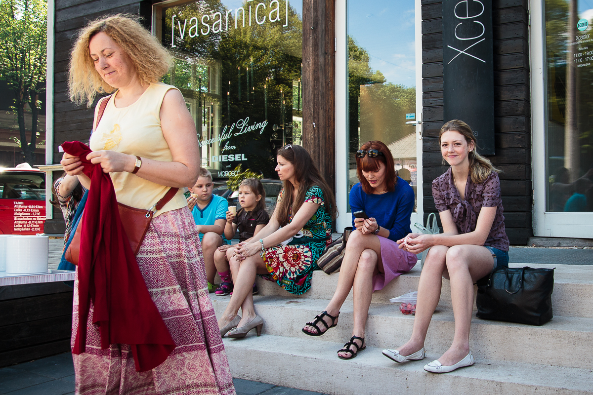 A woman with curly hair, wearing a white t-shirt and patterned skirt, holds a red garment while walking. Five people are sitting on stone steps: three children, an older woman in a floral dress, and a young woman in a purple top and skirt. They appear relaxed outside the Kalnciema kvartāla storefront with large windows on this modes diena.