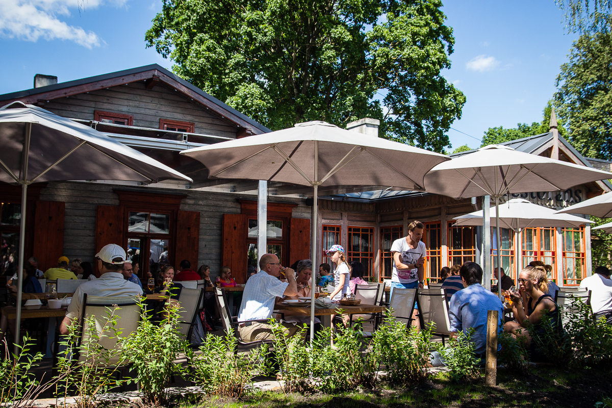 People seated at wooden tables under large patio umbrellas, enjoying meals and conversations at an outdoor cafe. The scene is vibrant, full of social activity. A rustic building with red-trimmed windows in Kalnciema kvartāla and lush green trees in the background complete the lively, sunny ambiance on this modes diena.
