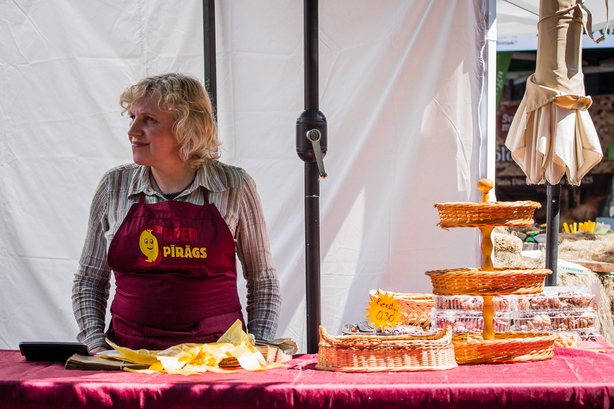 A person with light-colored, curly hair wearing a striped shirt and a red apron labeled "Rūder Pīrāgs" stands behind a booth at the Kalnciema kvartāla market. They are next to a tiered wicker stand holding baked goods. The booth has a maroon tablecloth, with yellow napkins and pastries on the table.