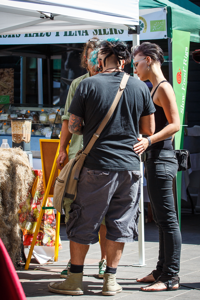 Three people, two men and one woman, stand closely together at an outdoor market stall during Kalnciema kvartāla modes diena. The woman, on the right, wears black clothing, and one man, with dreadlocks and cargo shorts, carries a shoulder bag. The other man is partially visible. Market tents and various products fill the background.
