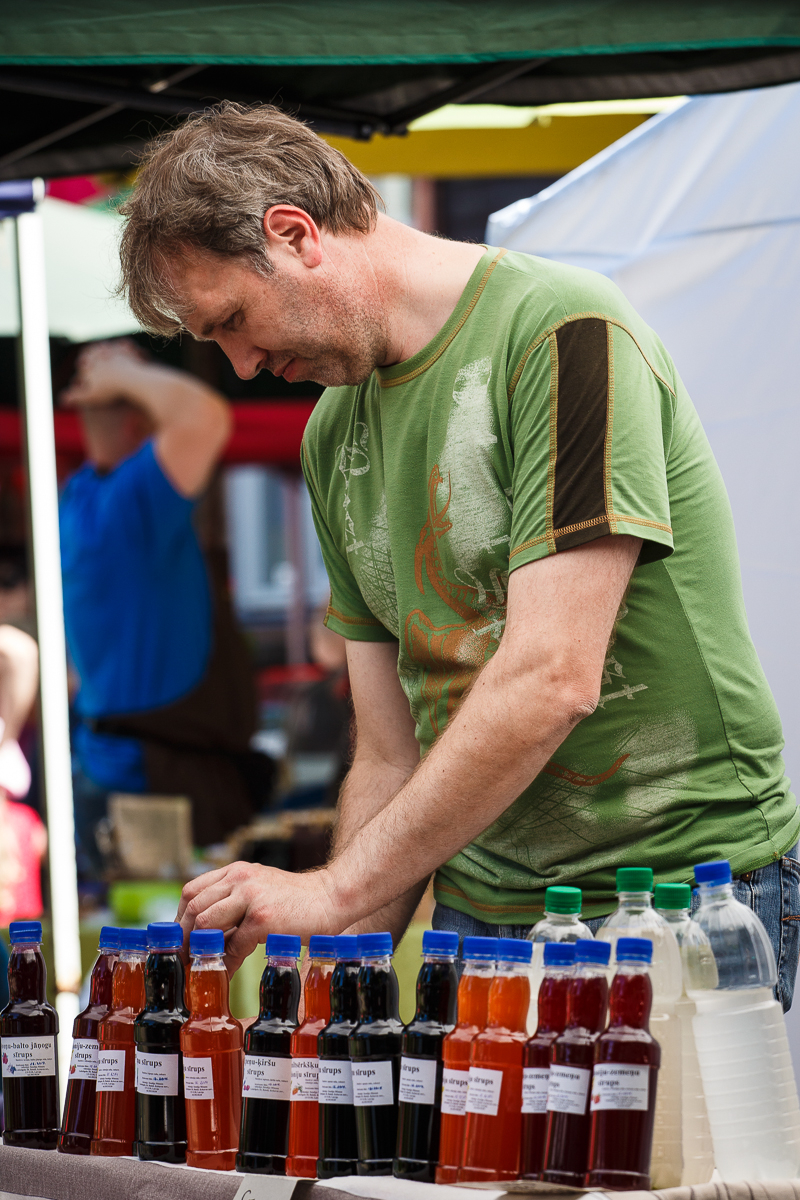 A vendor wearing a green T-shirt is standing at an outdoor market stall in Kalnciema kvartāls, arranging various colored bottles of juice or syrup. The bottles are neatly lined up on the table, each labeled with different flavors. The backdrop captures other market stalls and blurred people enjoying modes diena.