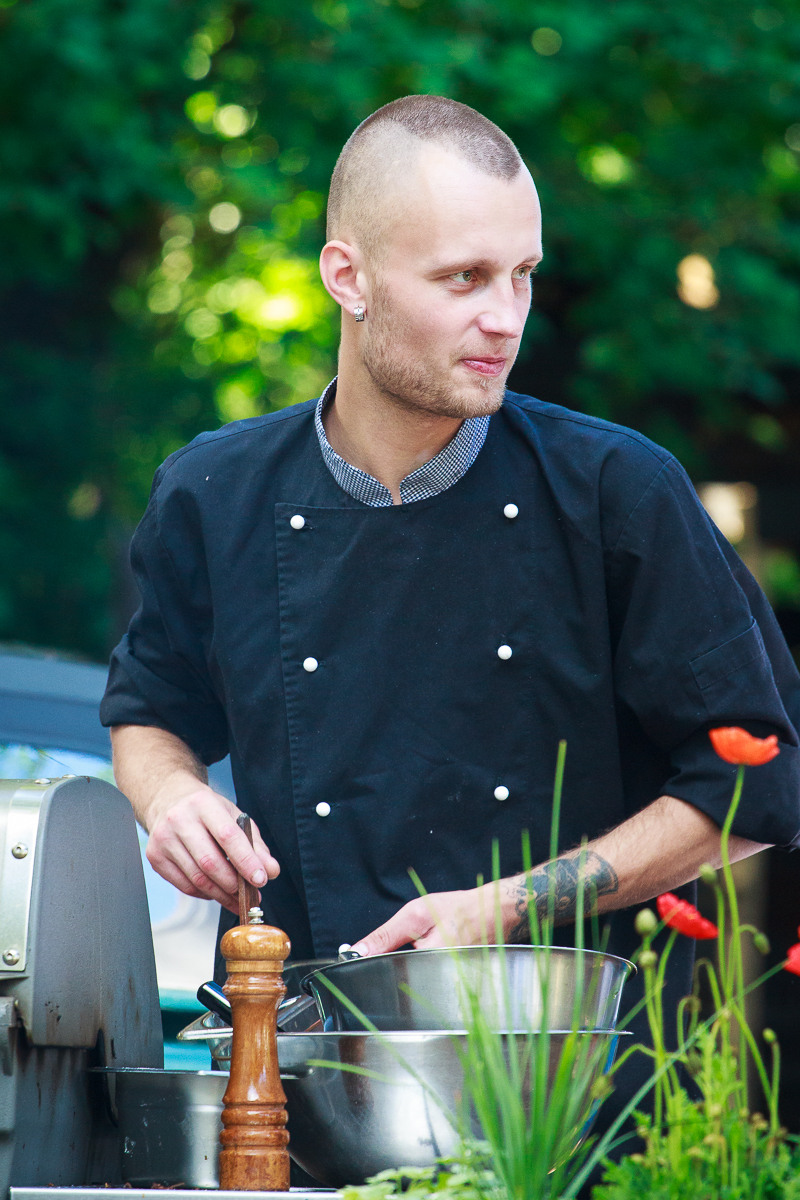 A chef with a shaved head and earrings, wearing a black double-breasted chef's coat, is standing outdoors at Kalnciema kvartāls preparing food. He holds a metal bowl and stirs its contents with a spoon, while a wooden pepper mill rests on the countertop beside him. Lush green trees are in the background.