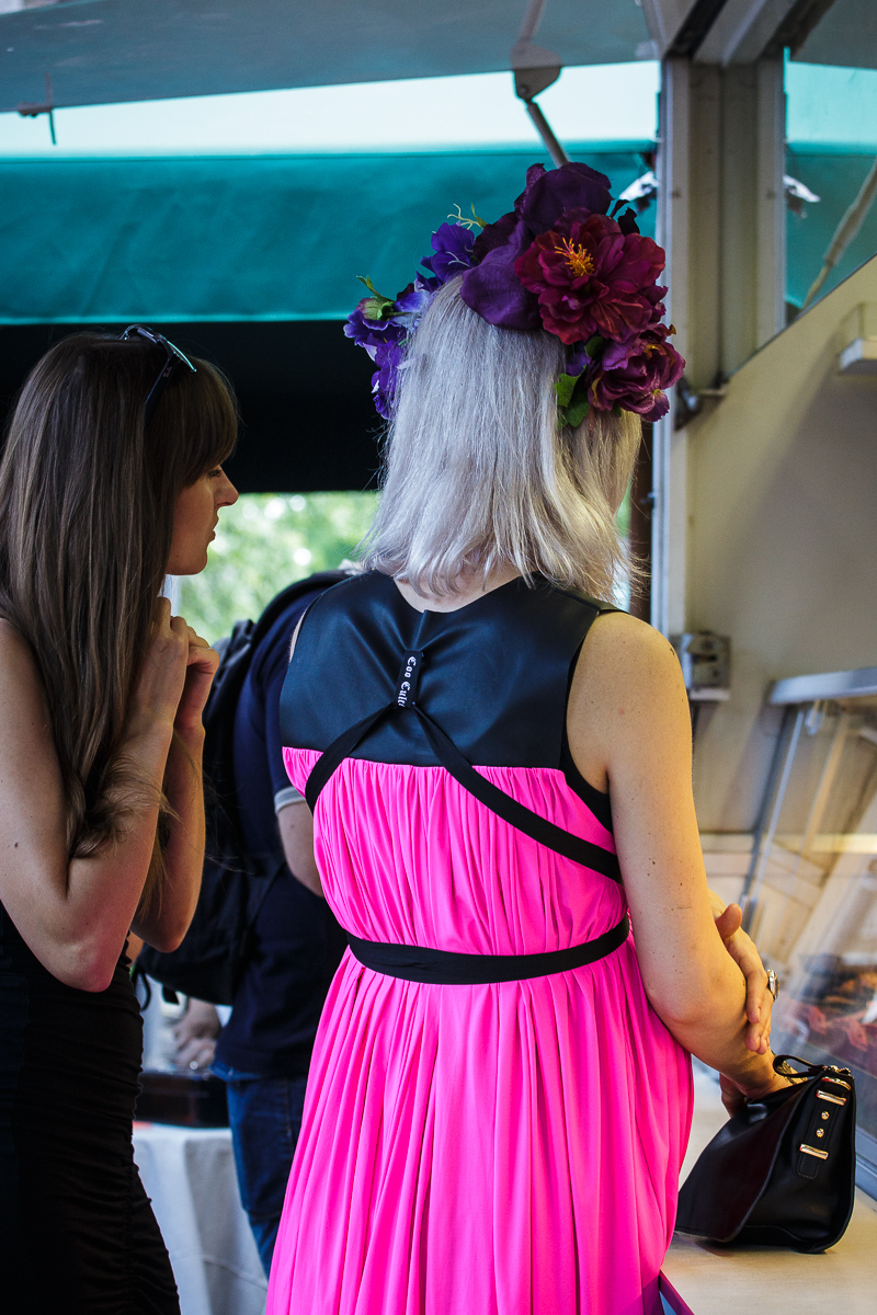 A woman with long, grey hair adorned with large, dark purple flowers stands at an outdoor market stall in Kalnciema kvartāls. She is wearing a bright pink dress with a black leather collar and belt. Another woman with long brown hair and sunglasses stands beside her, looking at the Modes Diena stall.
