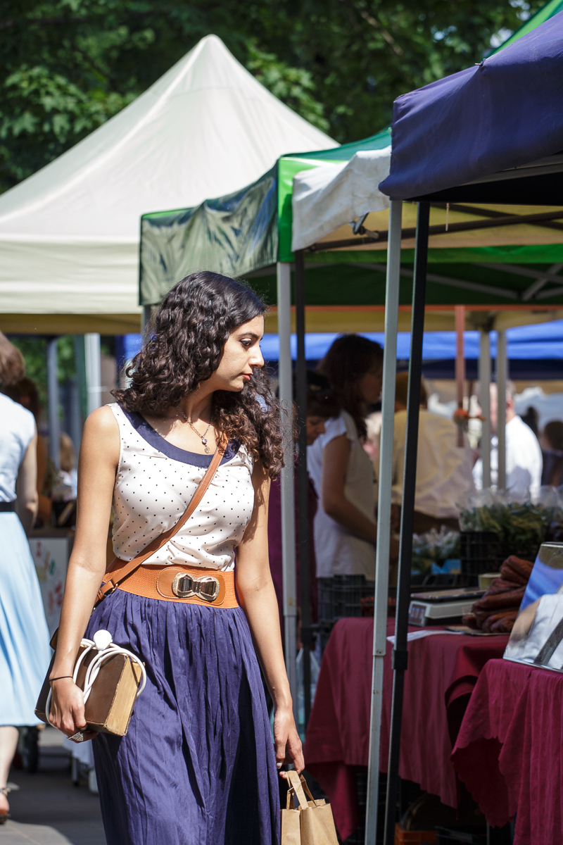 A woman with long curly hair, wearing a polka dot sleeveless top and a navy blue skirt with a wide belt, holds a brown paper bag in each hand as she looks to her left. She stands in front of vendor stalls at Kalnciema kvartāla modes diena, with canopy tents providing shade and people browsing in the background.