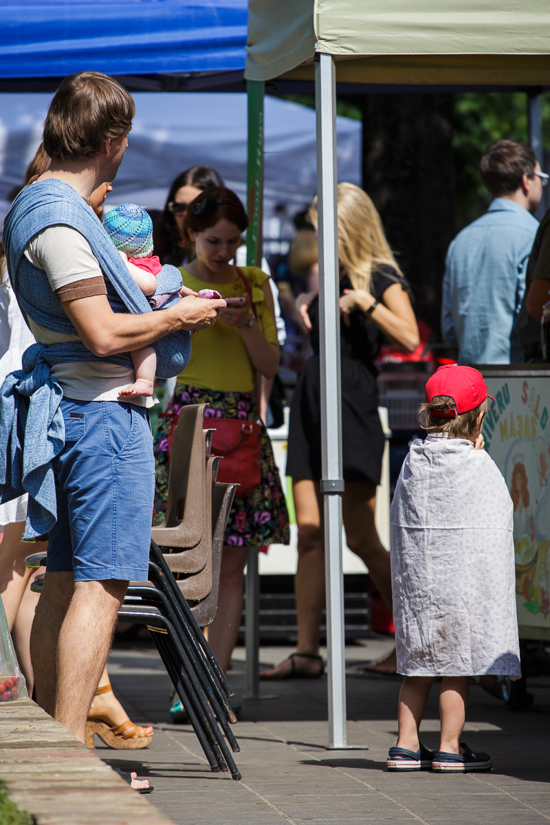 Adults and children enjoy the festivities at a Kalnciema kvartāla event. A man in a blue shirt, blue shorts, and sandals holds a baby in a floral hat and blue wrap. A boy in a red cap and white cape stands near a sign as people in colorful clothing gather under blue and yellow tents in the background.