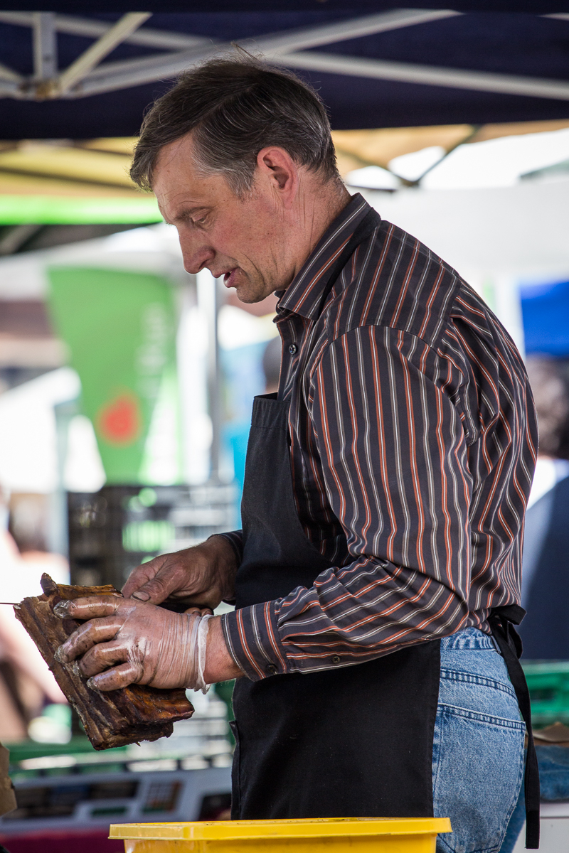 A man, wearing a black apron over a striped shirt and jeans, is working at Kalnciema kvartāls market. He is handling a rack of ribs while wearing plastic gloves. The background features market stalls with green and blue canopies and blurred patrons on modes diena.