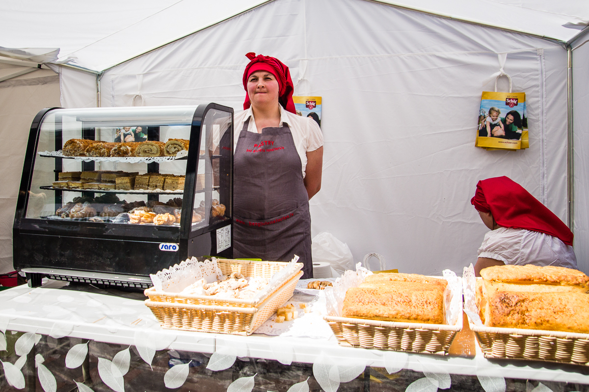 Two women are in a white tent bakery stand at the Kalnciema kvartāla market. One woman stands behind a display case filled with various pastries while wearing a red headscarf and gray apron. The other woman, also in a red headscarf, attends to something on the side. Baskets of bread and pastries are on the table in front.