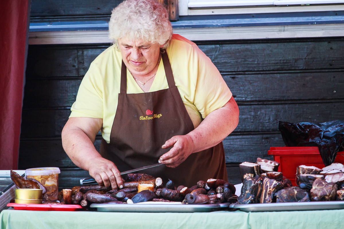 A woman with curly blonde hair, wearing a yellow shirt and a brown apron, prepares assortments of sausages and meat at Kalnciema kvartāla's wooden counter during modes diena. Various meats and containers are spread out in front of her. The background showcases a wooden wall and a red plastic container by her side.