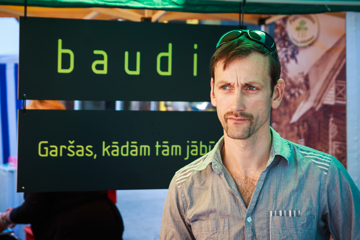 A man with short brown hair and sunglasses resting on his head stands in front of a tented booth at Kalnciema kvartāls. The sign behind him reads "baudi" and "Garšas, kādām tām jābūt." He is wearing a blue button-up shirt and appears to be looking slightly to the side, enjoying the modes diena atmosphere.