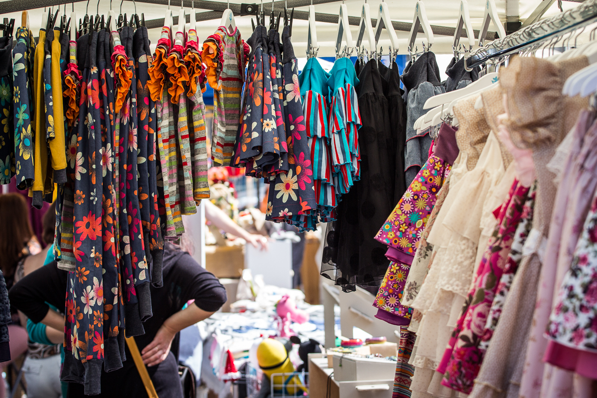A colorful market stall at Kalnciema kvartāla displays an array of children's clothing. On the left side, rows of bright floral and striped dresses hang neatly, while the right side shows pastel-toned, lacy dresses. In the background, shoppers peruse more items under a white canopy on this lively modes diena.