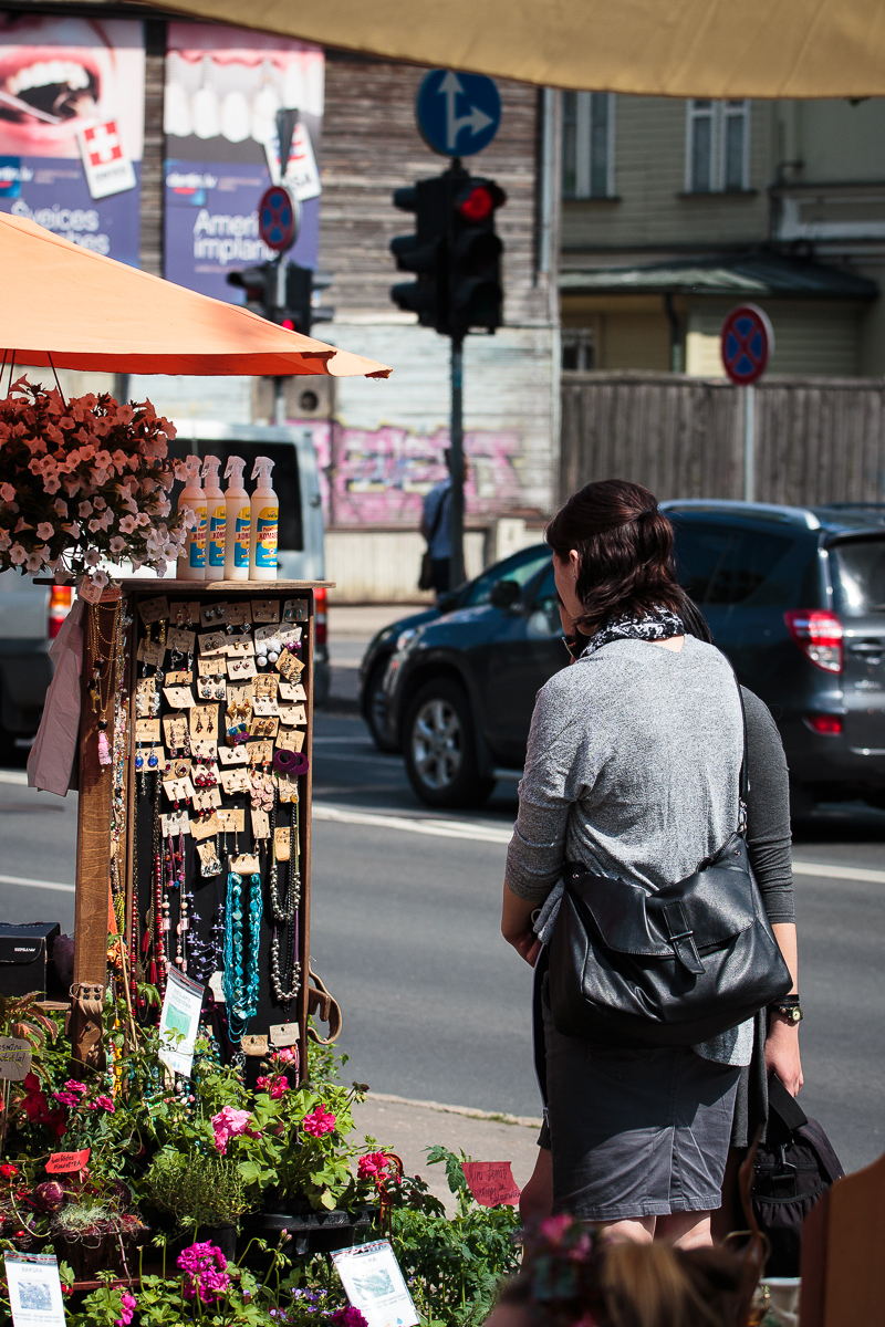 A person with dark hair and a gray top is standing in front of an outdoor market stall adorned with various jewelry items such as necklaces and earrings. The stall, part of Kalnciema kvartāla's lively scene, is shaded by an orange umbrella, with flowers at its base. A street with cars and traffic signals is in the background on this modes diena.