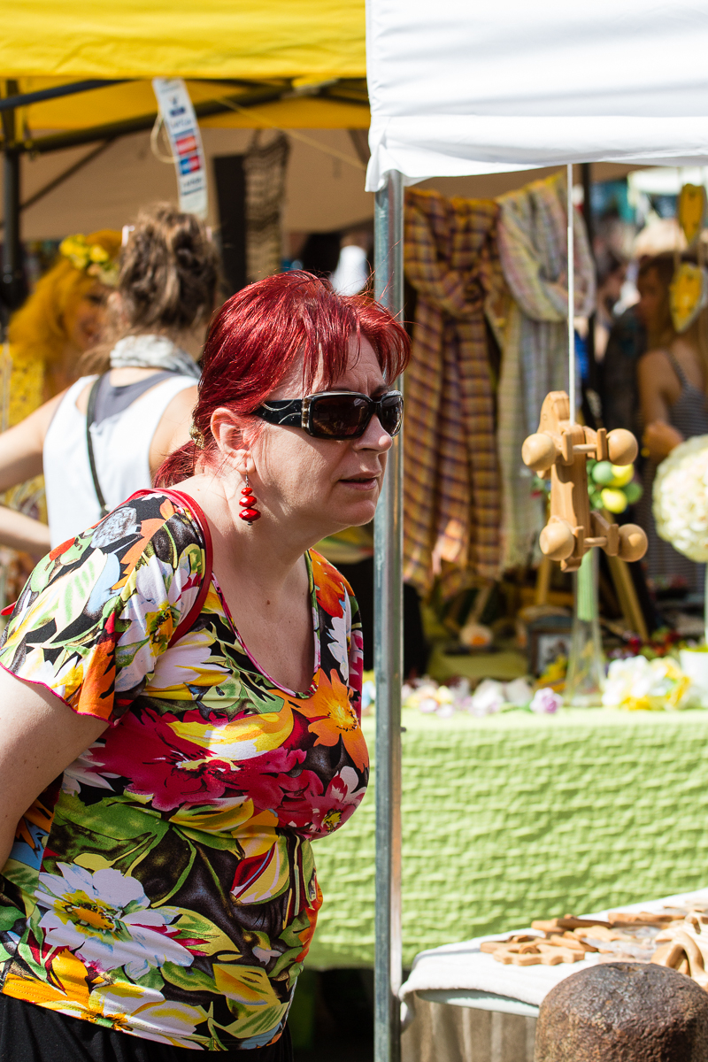 A woman with red hair, wearing sunglasses, a bright floral shirt, and red earrings is browsing items at the Kalnciema kvartāla market during the modes diena event. Behind her are stalls with colorful fabric and various goods on display. The background also shows other shoppers and a yellow canopy.