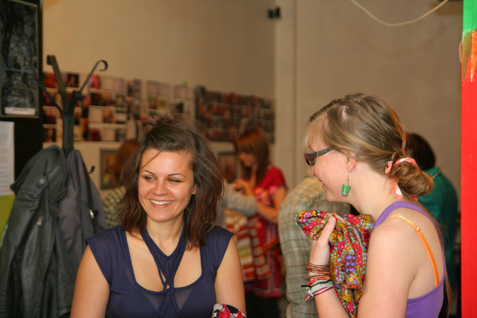Two women are engaged in talka in a warmly lit indoor setting. The woman on the left, smiling, has shoulder-length brown hair and wears a sleeveless navy blue top. The woman on the right has blonde hair, wears glasses, and holds colorful fabric. They are among other people and some wall decor.
