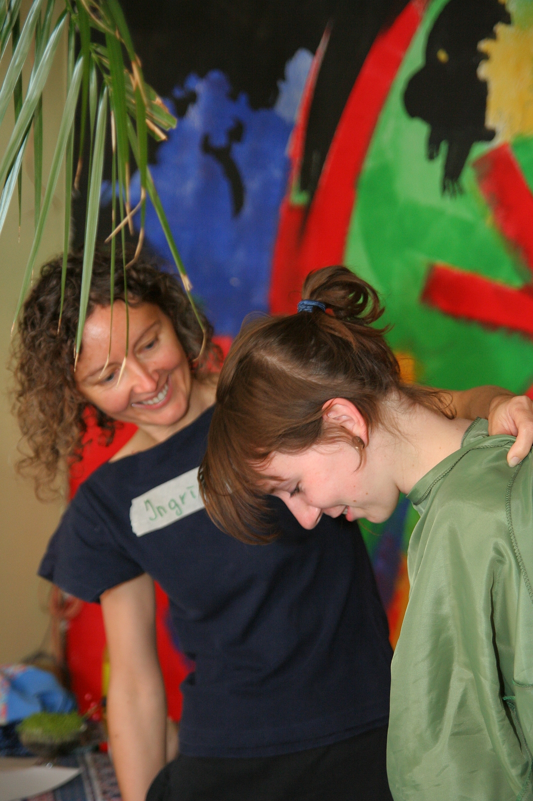 A person with curly hair and a shirt that has a name tag is smiling and leaning toward another person with brown hair, who is laughing and looking down. They are standing in front of a colorful abstract mural, immersed in their modes of communication. Some green leaves hang down from the top of the image.