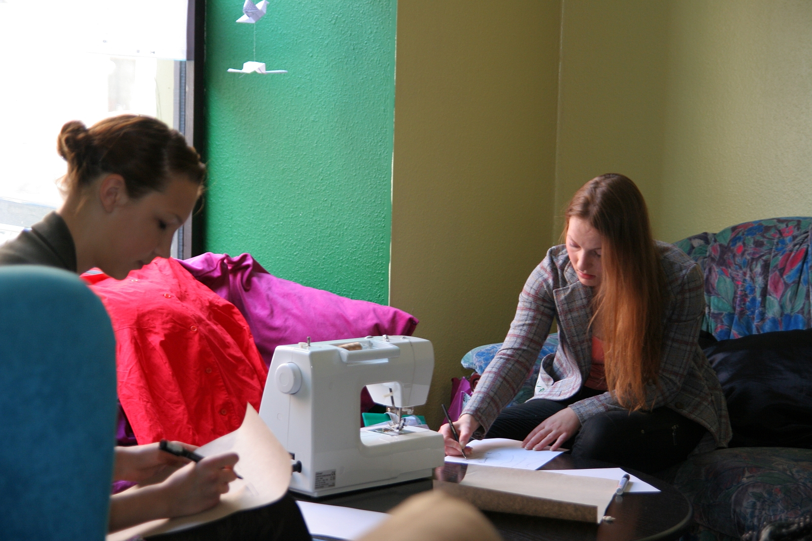 Two women focus on their work inside a room with green and yellow walls. One woman writes notes at a desk with a sewing machine and fabric, while the other, seated nearby with a notebook, writes intently. They sit on a couch and chair, respectively, near a window letting in natural light—modes talka filling the air.