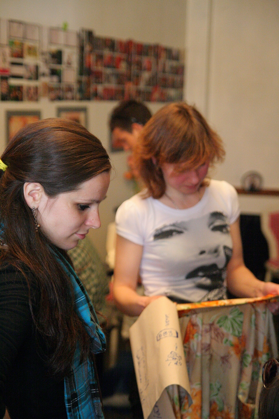 Two women are engaged in an activity indoors. The woman on the left, with long dark hair and a turquoise scarf, seems to be in deep modes talka with her friend. The woman on the right, wearing a face-printed white T-shirt and holding a beige patterned cloth, listens intently. Blurred images adorn the background walls.
