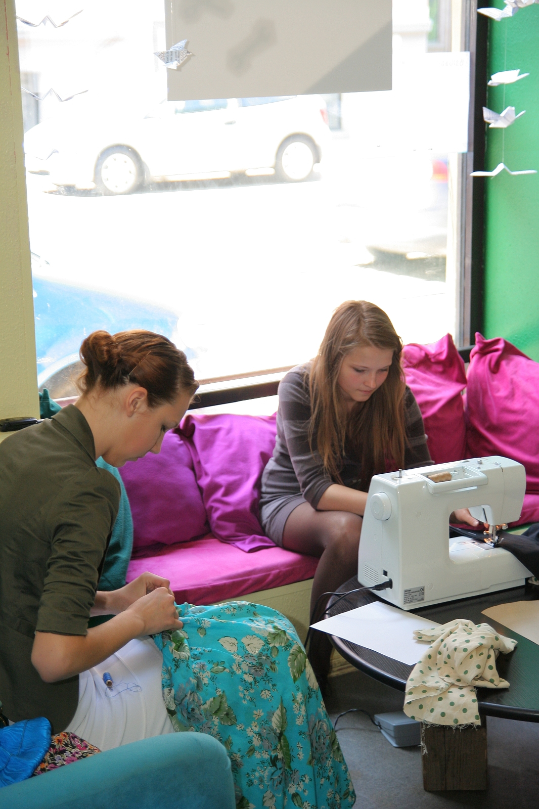 Two women are engaged in sewing activities in a bright room with large windows. One woman on the left hand-sews fabric adorned in blue patterns, while the woman on the right uses a white Modes sewing machine. The cozy room features pink and purple cushions, with paper cranes hanging in the window.