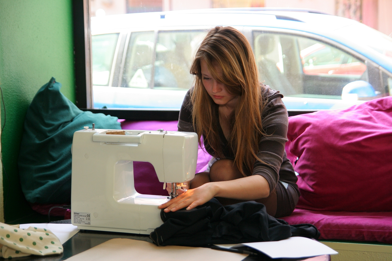 A woman with long hair is using a Modes talka sewing machine while seated on a magenta cushioned bench next to a green wall. She is focused on stitching fabric. There are papers and more fabric around her. A large window behind her shows parked cars, providing natural light to the workspace.