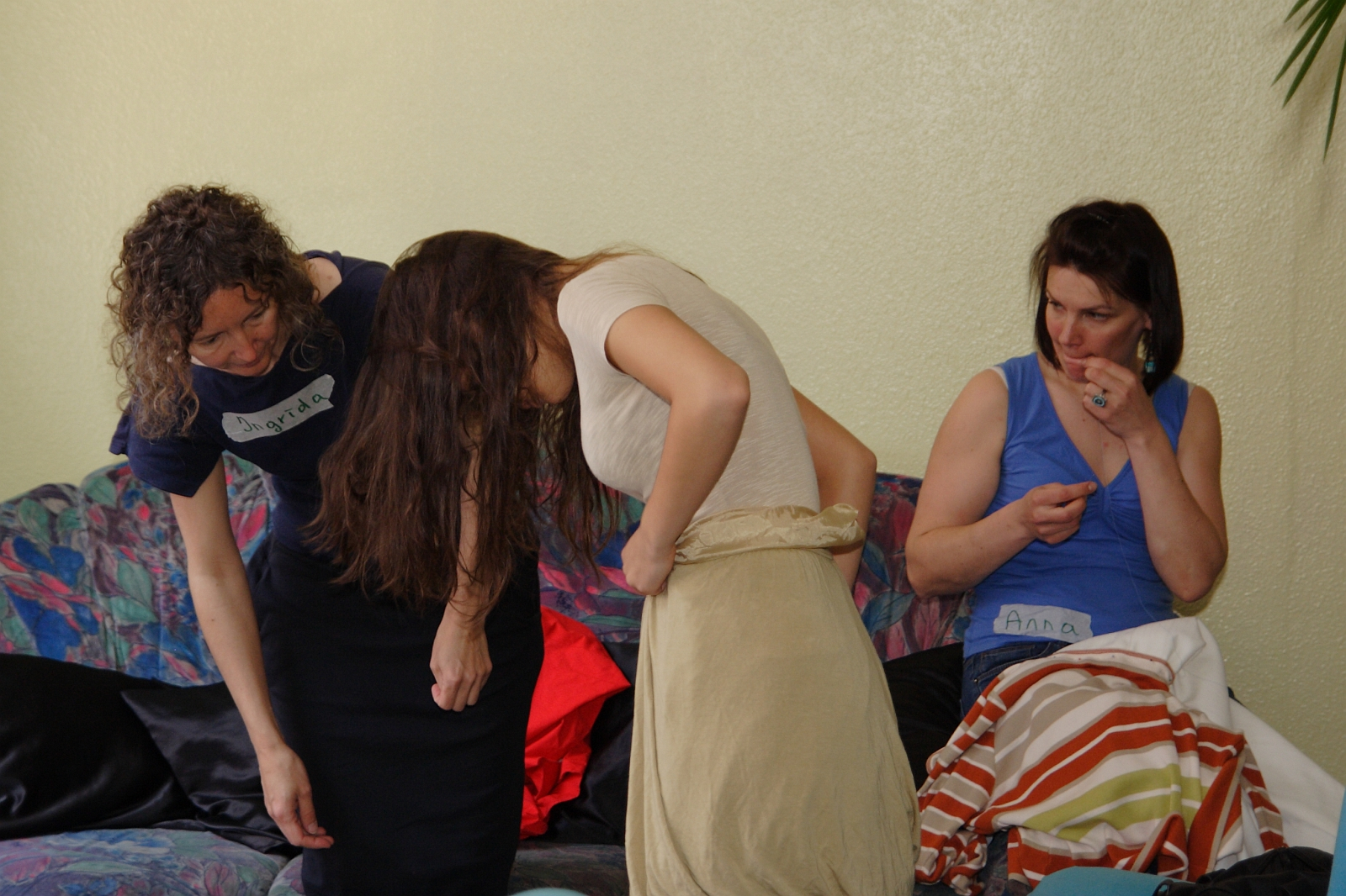 Three women are seated on a colorful couch. Two women on the left are bending over, examining something on the floor. The woman on the far left has curly hair and a blue top, and the middle woman has long hair and a beige skirt. The third woman on the right is sitting upright in a blue top, seemingly lost in talka modes of thought.