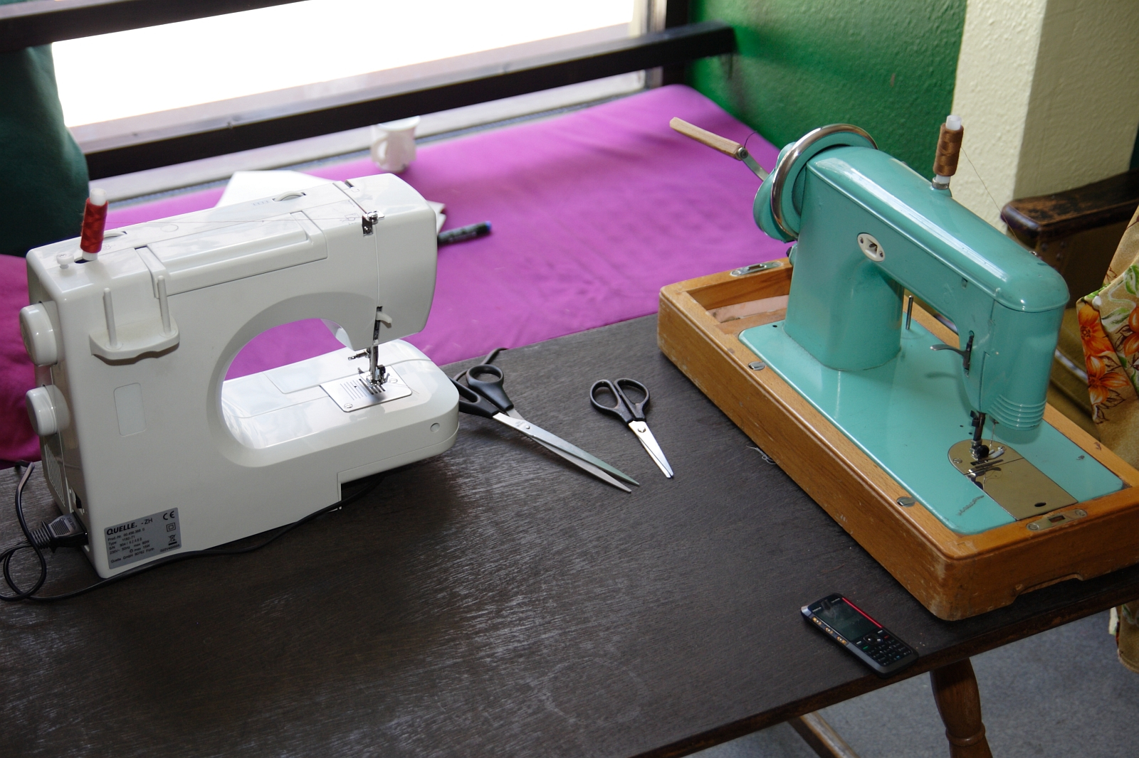 Two sewing machines on a wooden table: a modern white electric machine on the left and a vintage teal manual machine on the right. Scissors, thread, and a black mobile phone are also on the table, perfect for modes talka enthusiasts. A purple cloth and window are in the background.