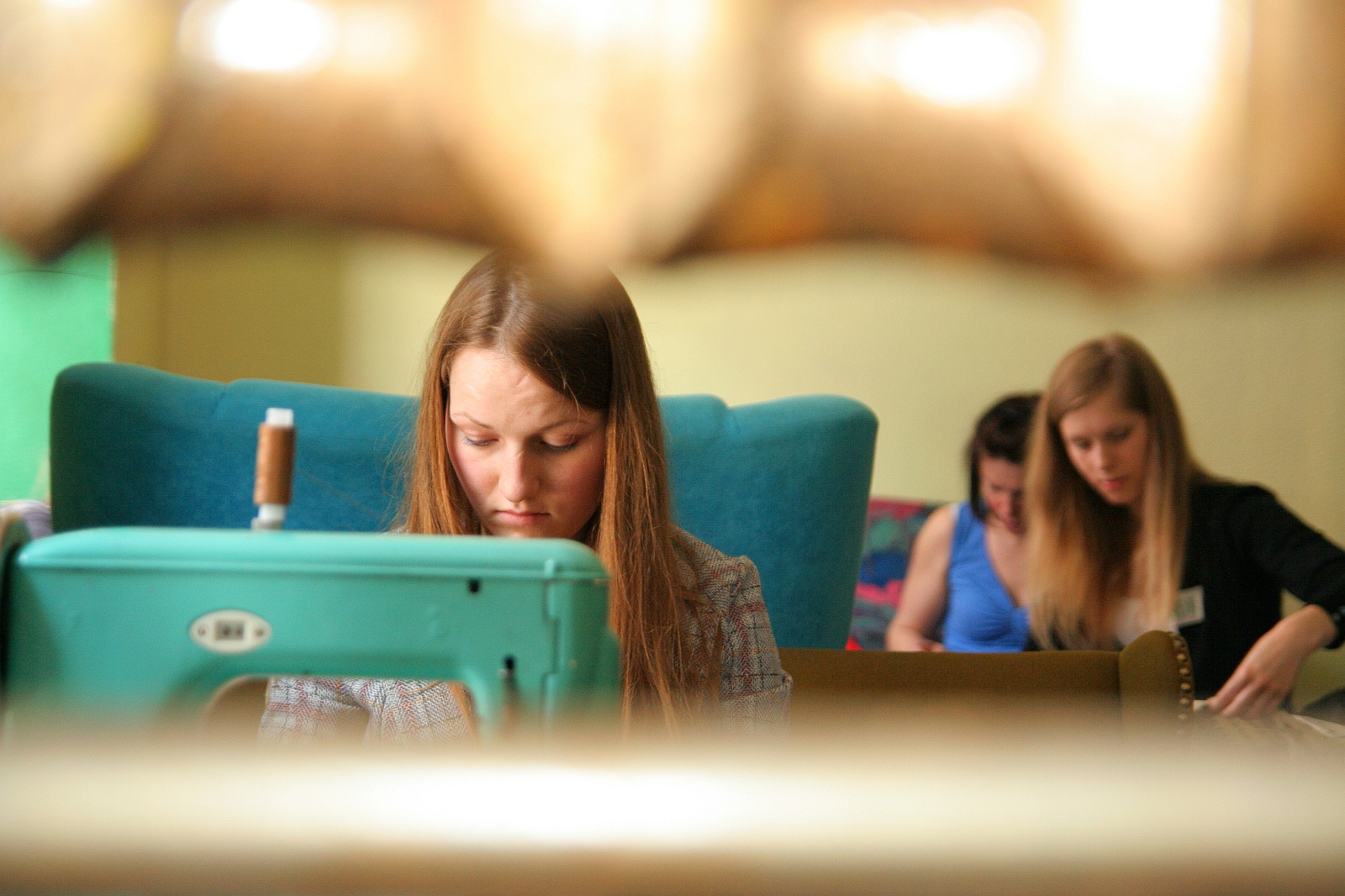 A young woman with long brown hair is focused on sewing with a turquoise machine in the foreground. Behind her, two other young women are engaged in talka on a couch. The scene is warmly lit, giving a cozy and intimate atmosphere, capturing their creative modes perfectly.