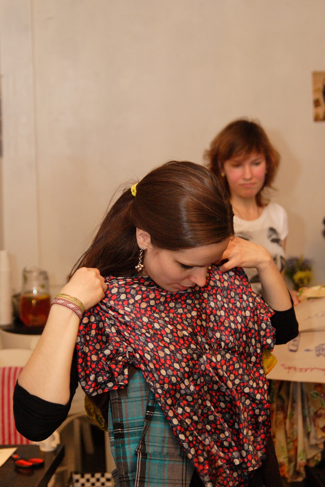 A woman with long brown hair, wearing a plaid shirt, holds up a red and blue patterned shirt in front of her, inspecting it closely. Another woman with short brown hair, wearing a light gray top, stands in the background observing. The setting appears to be indoors with various items around, creating a cozy Modes talka atmosphere.