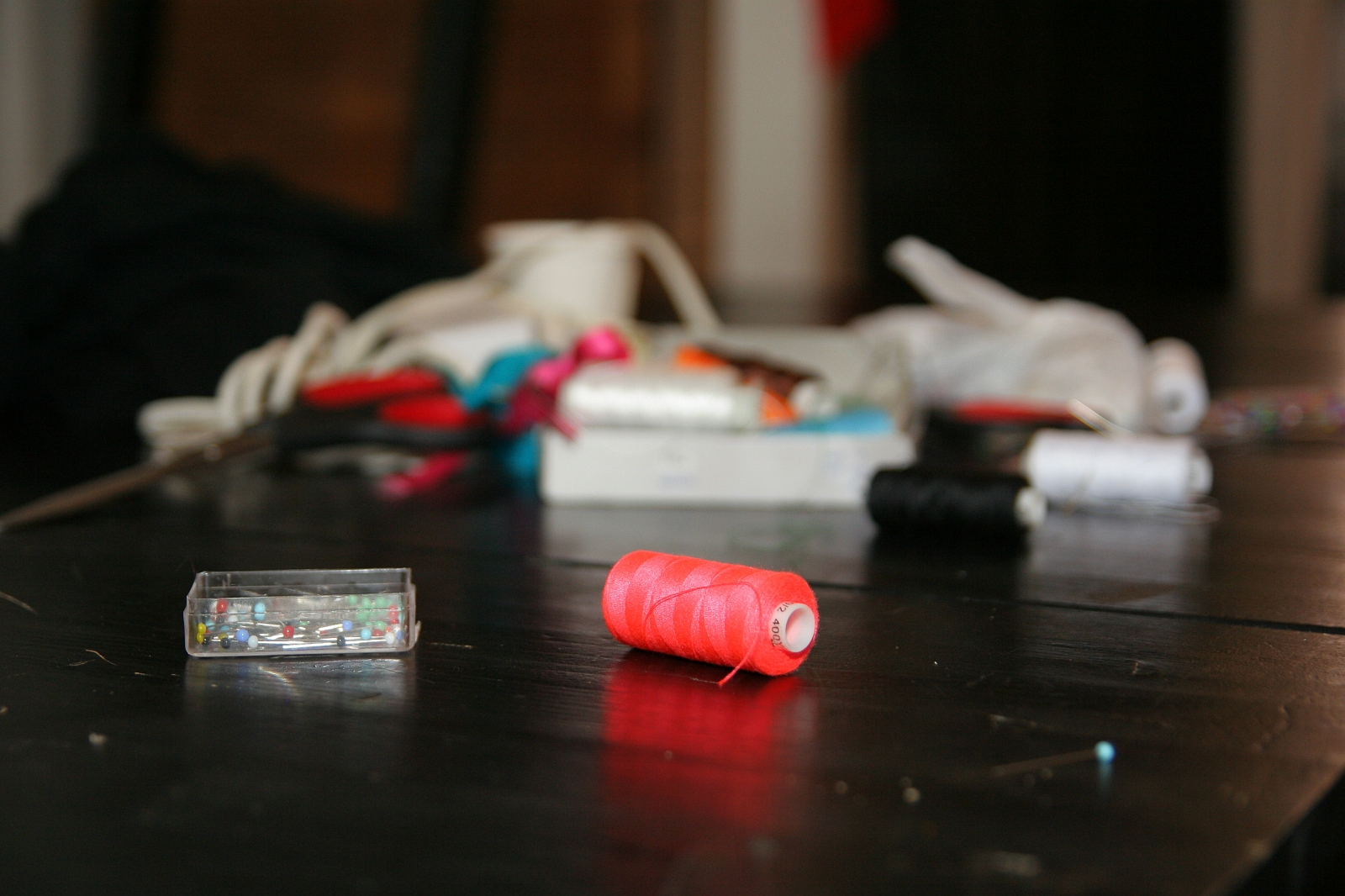 A close-up of a spool of red thread on a dark wooden table. In the background, the table is scattered with sewing supplies, including a box of multicolored pins, various spools of thread, fabric scraps, and an assortment of sewing notions—all slightly blurred to focus on the talka red thread spool.
