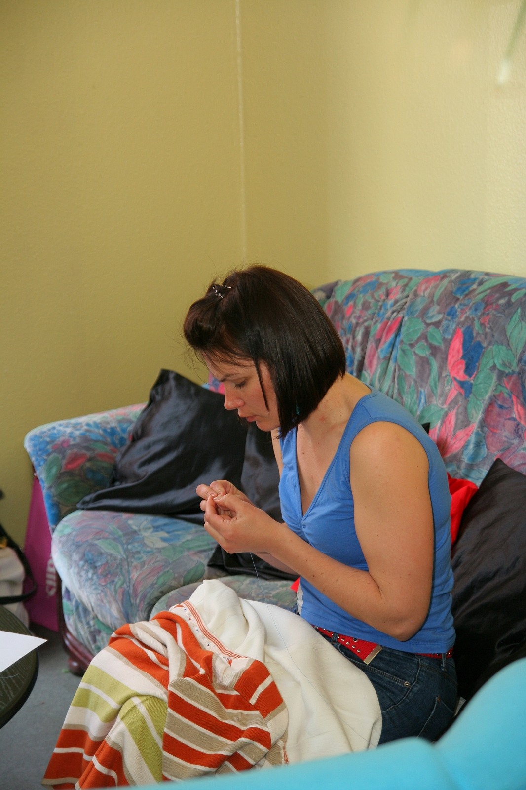 A woman with straight, shoulder-length hair and wearing a blue sleeveless top is sewing a white and orange striped fabric. Seated on a colorful floral-patterned couch with various cushions, in a room with light-yellow walls, she appears focused on her task, engrossed in her modes talka.