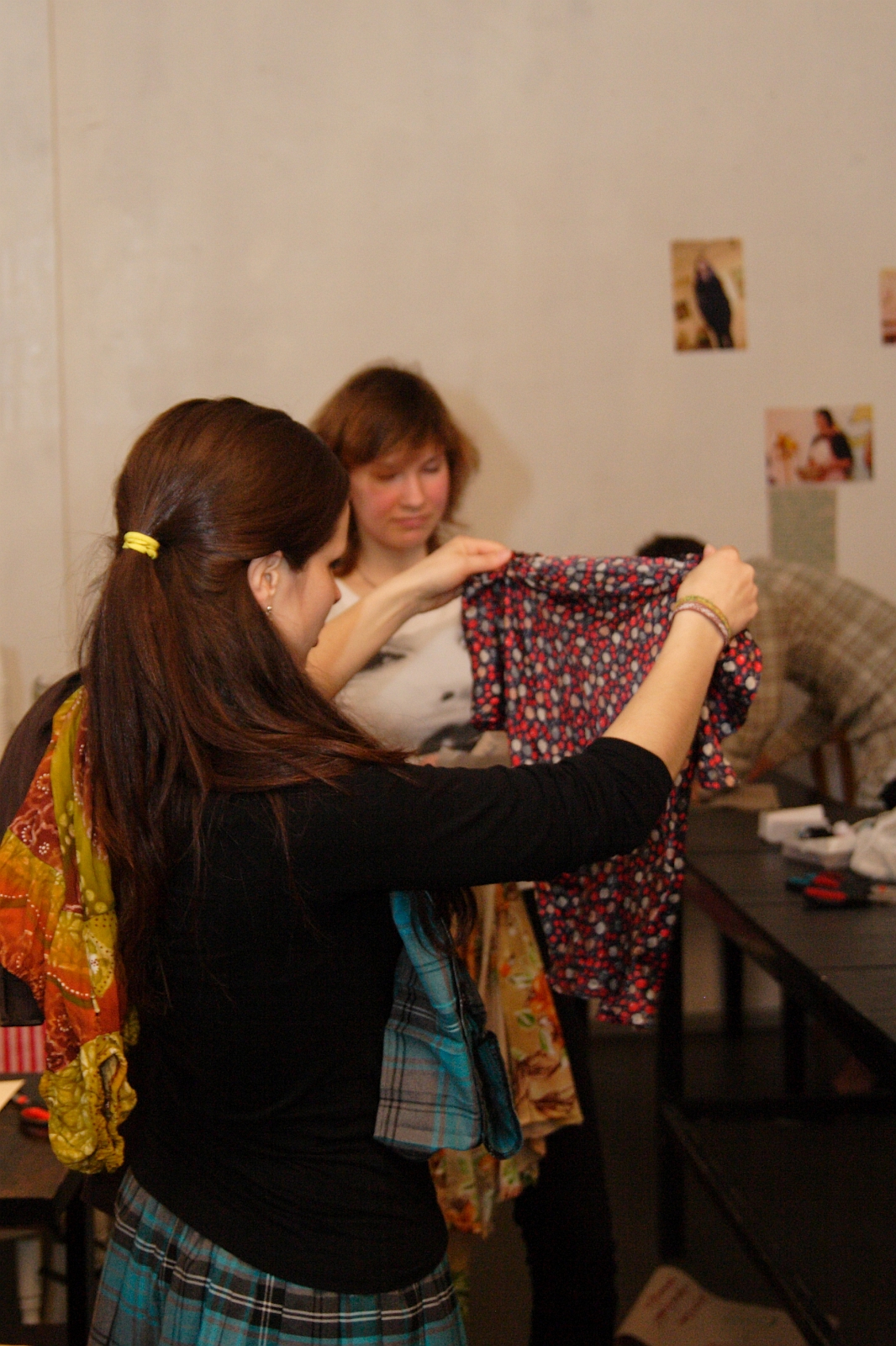 Two women hold and examine pieces of clothing in a room with light-colored walls and various pictures pinned to them. The foreground woman, sporting a colorful scarf, exudes Modes talka elegance. The background woman with shoulder-length brown hair holds up a patterned shirt.