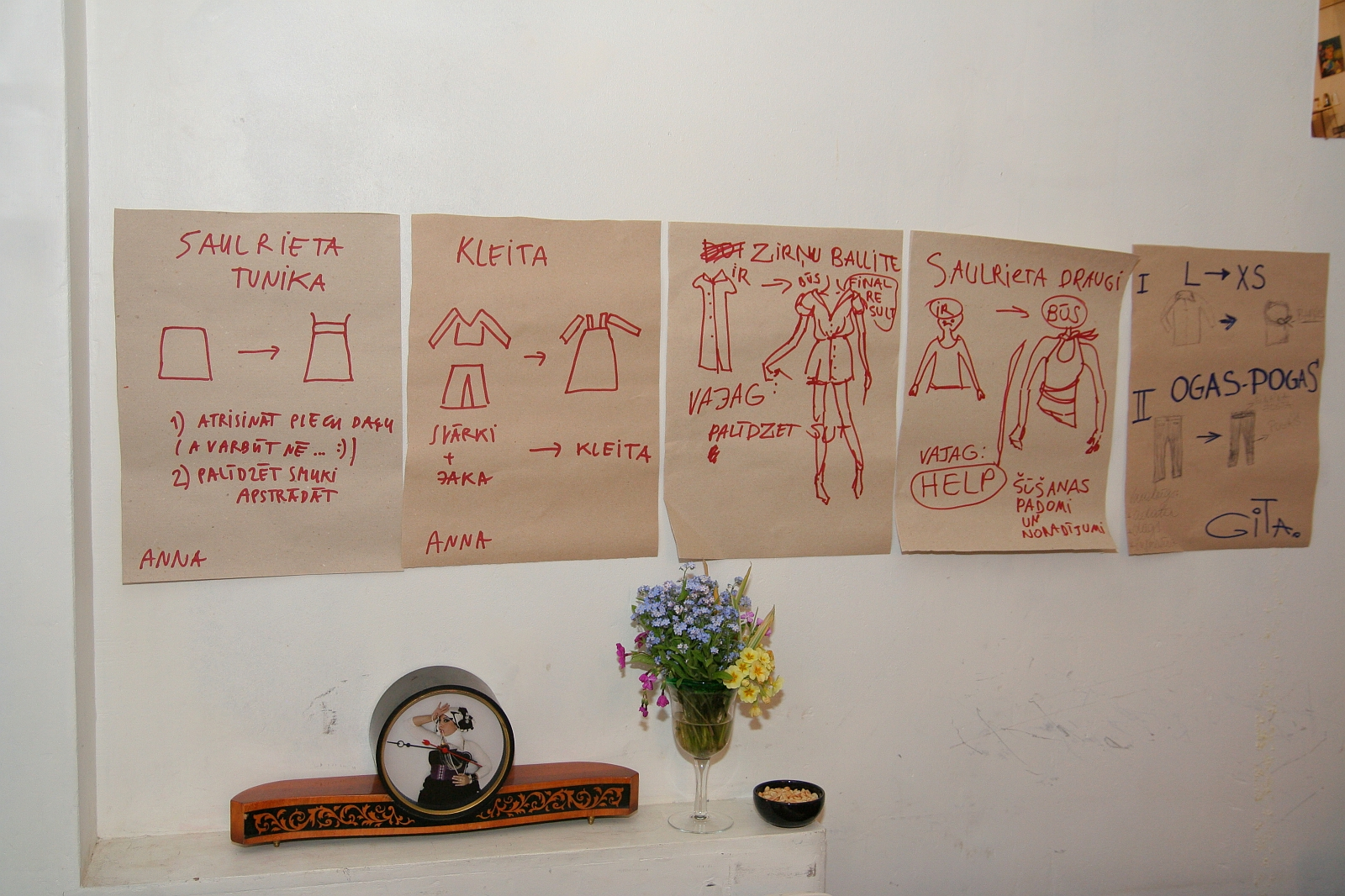 Five hand-drawn instructional posters featuring step-by-step illustrations on how to make various clothing items in different Modes are displayed on a white wall. Below the posters, a small shelf holds a clock, a potted plant with colorful flowers, and a glass jar of talka.