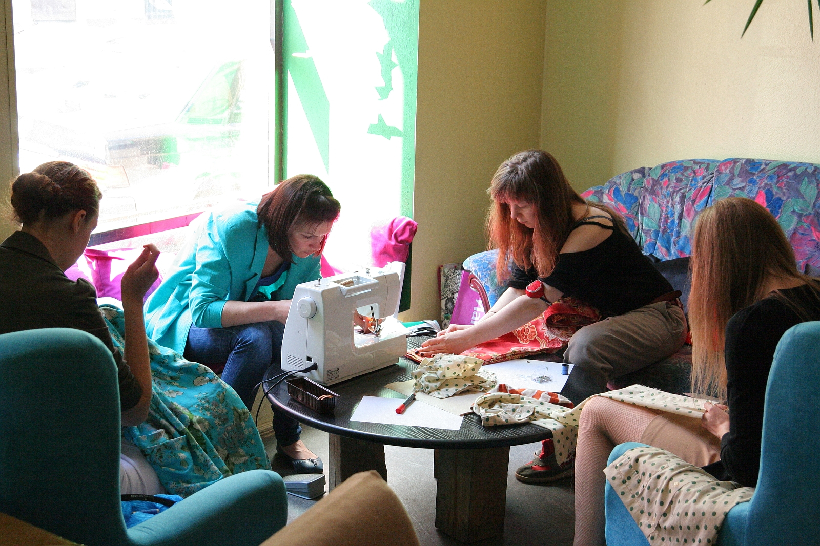 Four women are engaged in sewing activities in a cozy room. Two women are using a sewing machine on a round table, while another two are hand-sewing. They are surrounded by colorful fabrics and seated on comfortable chairs near a window, creating a relaxed, collaborative Modes Talka atmosphere.