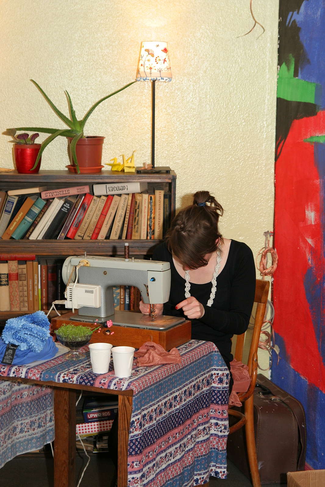 A woman sits at a wooden table using a sewing machine, surrounded by various fabrics and cups. The table is covered with a colorful, patterned cloth. Engaging in modes talka, she finds inspiration from the bookshelf filled with books and potted plants behind her. A vibrant, abstract painting decorates the wall as a lamp illuminates the space.