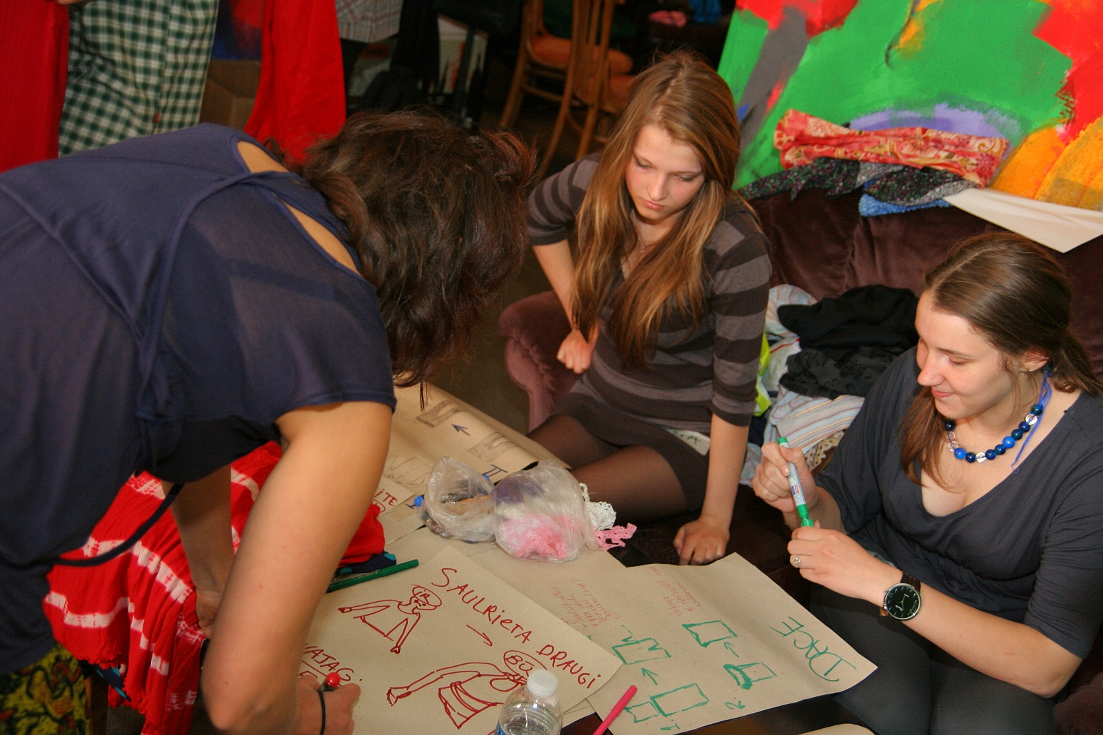 Three women are gathered around a table, engaging in collaborative drawing, or "talka." They are using markers on large sheets of paper filled with sketches of clothing. The scene is set in a colorful, busy environment with various fabrics and artworks illustrating different fashion modes in the background.