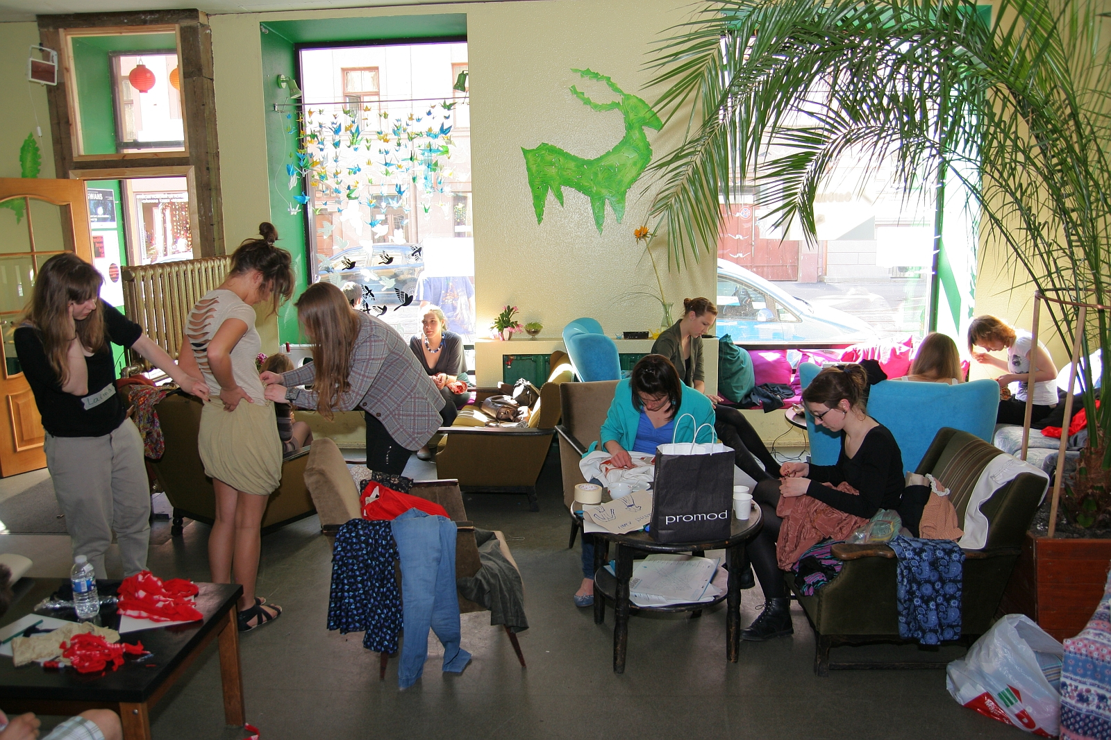 A lively room filled with people engaged in various activities. Some are standing, others are seated, all focused on tasks like sewing, drawing, or crafting using Modes talka techniques. The walls are adorned with green unicorn-themed decorations. A large potted plant and windows allow natural light to brighten the space.