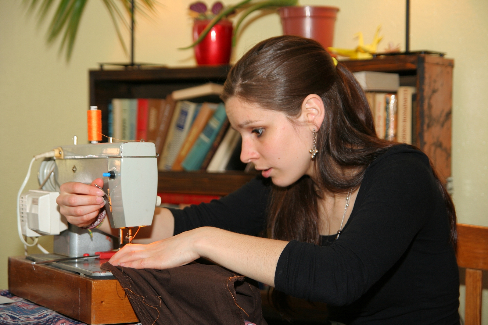A young woman with long brown hair is using a light-colored sewing machine. She is focused, guiding brown fabric under the needle with one hand while adjusting settings using the various Modes with the other. The scene is indoors, with bookshelves and potted plants in the background, creating a cozy, lived-in feel.